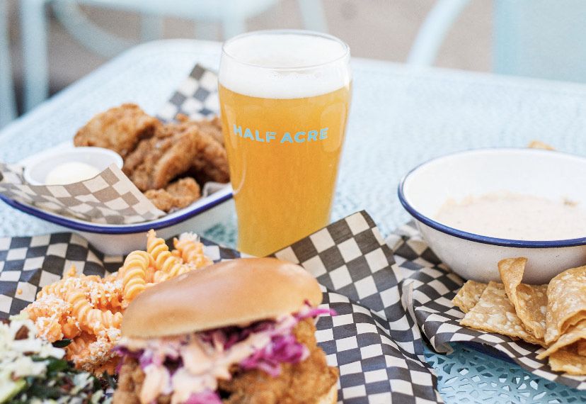 A table laid with sandwiches and sides in baskets and a full glass of beer that reads “Half Acre.”