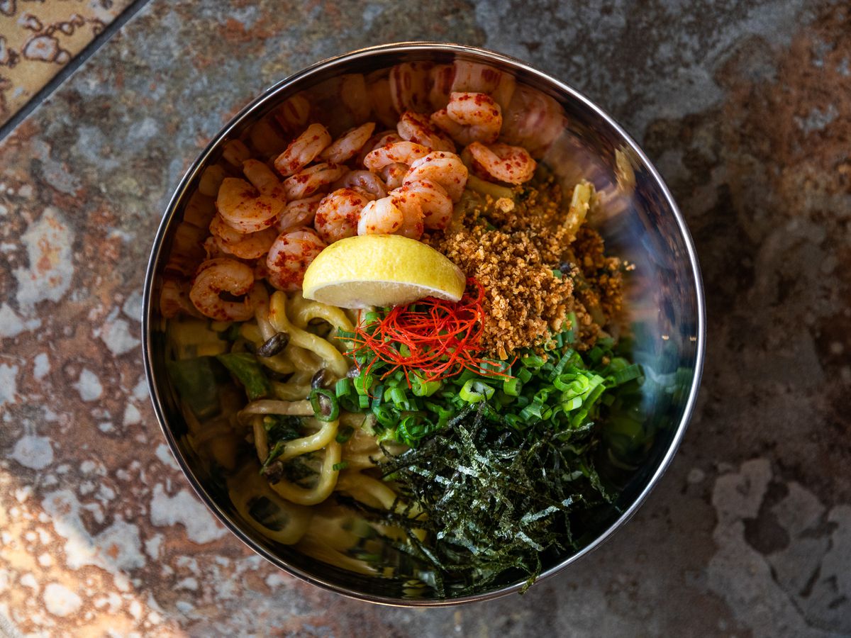 Butter udon noodles with poach shrimp, a lemon wedge, and herbs in a metal bowl. 