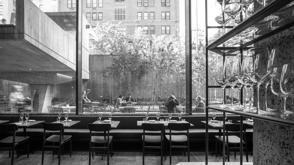 A black-and-white photo of a dining room, facing floor-to-ceiling windows, with shelves of wine glasses in the foreground