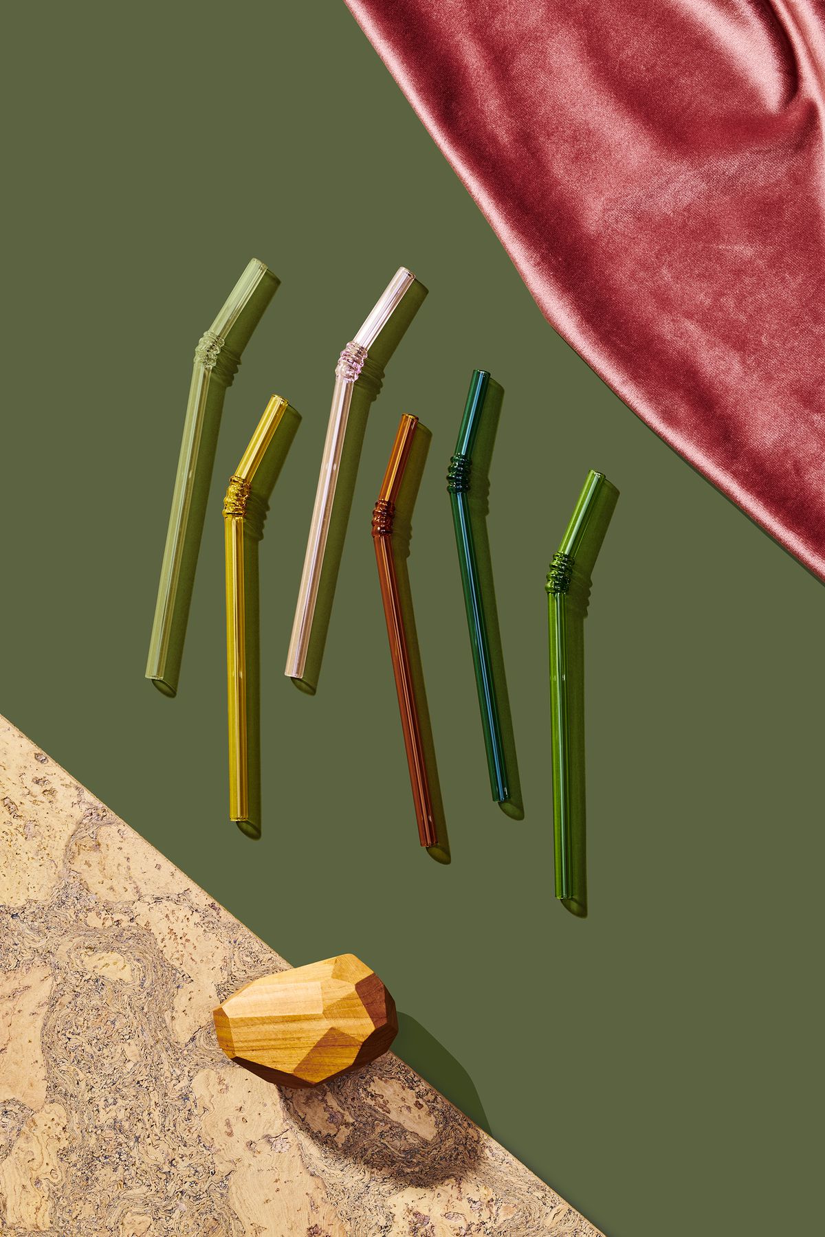A group of six multicolored glass straws which are part of the 2019 Holiday Gift Guide for Curbed. The straws are on a flat green surface and are flanked by pink fabric and various design objects.
