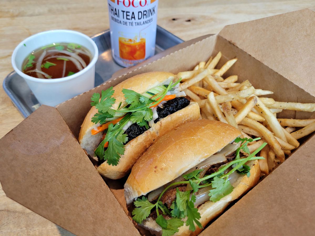 A pair of banh mi in a box with French fries.