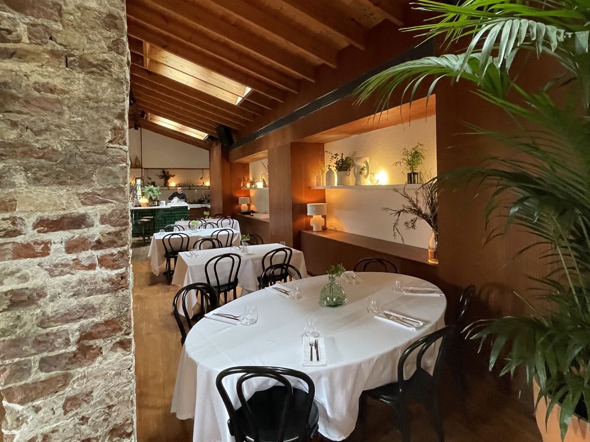 A restaurant interior with white tablecloth-set tables beneath an angled roof studded with skylights. Nearby there’s an old interior brick wall and mellow light and plant indentations in another wood-lined wall.