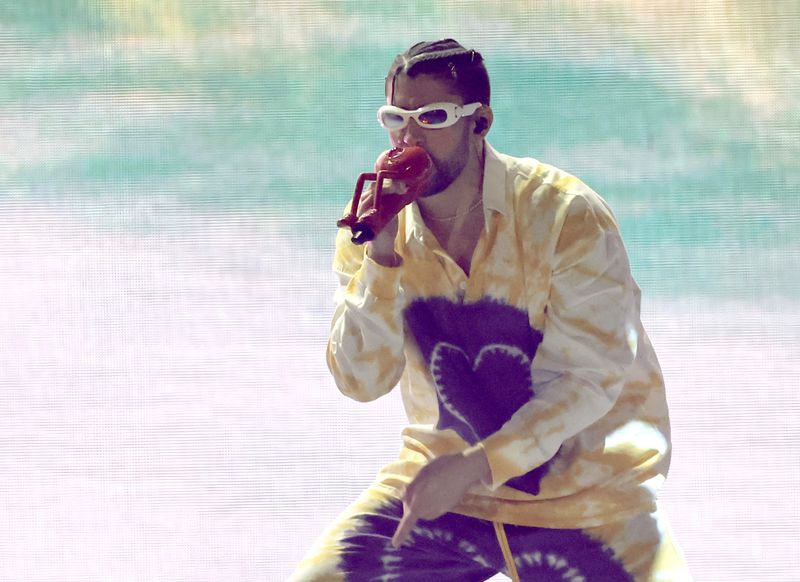 Bad Bunny, wearing large white-framed sunglasses and a patterned yellow outfit with large purple hearts, sings into a microphone. 