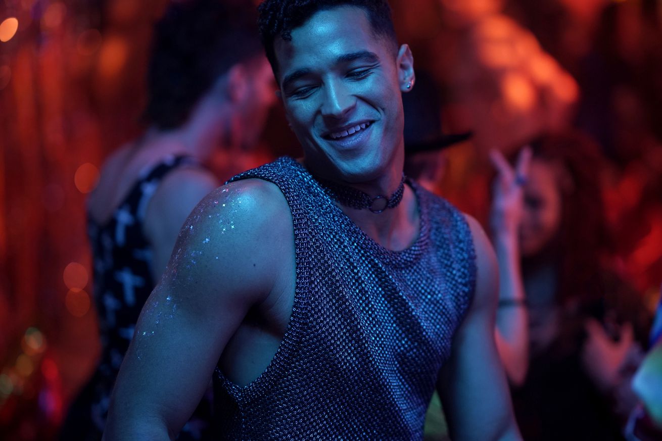 A man in a chainmail tank top dancing in a dimly-lit club alongside other partygoers.