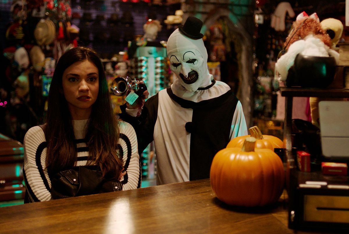 A young woman standing at the counter of a Halloween decoration store with a leering man in black and white clown makeup squeaking a horn in her ear.