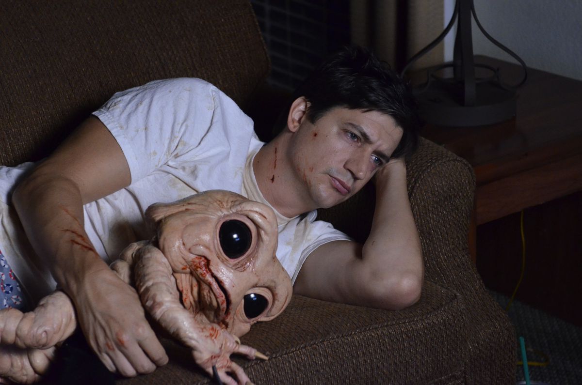 Duncan (Ken Marino) se acuesta en el sofá con su camiseta blanca sosteniendo a Milo, un demonio trasero que se parece vagamente al bebé de Dinosaurios pero con ojos gigantes de Baby Yoda.