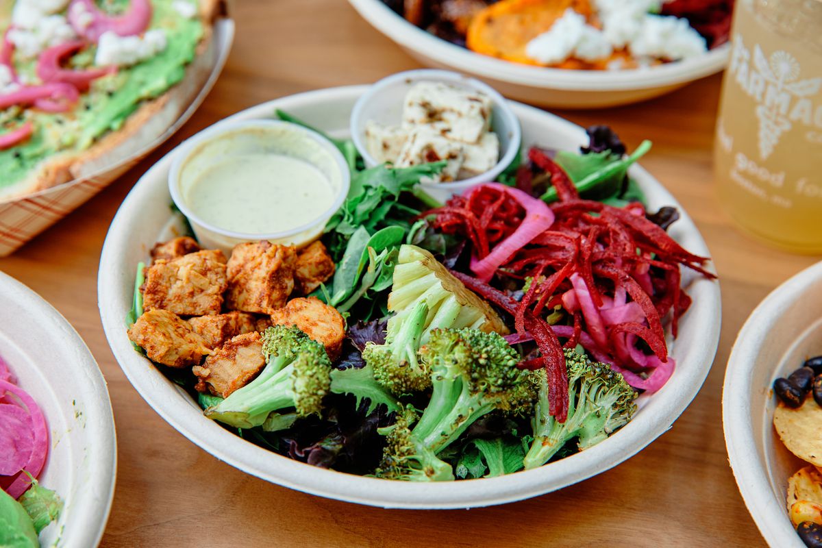 A paper takeout bowl is filled with a salad of broccoli, greens, thinly sliced beets, chicken, and more.