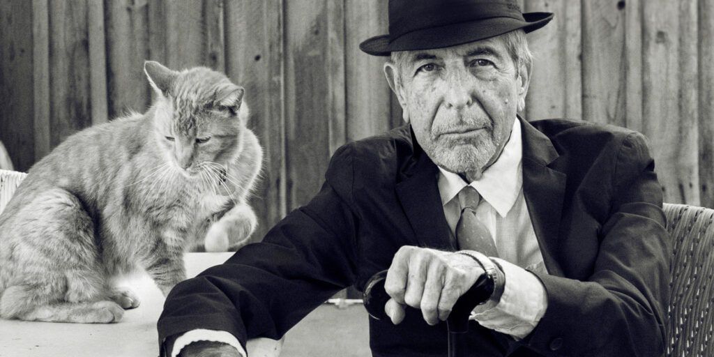 A black and white archival photo of Leonard Cohen in a dark suit sitting in a chair holding a cane next to a table with a cat on it.