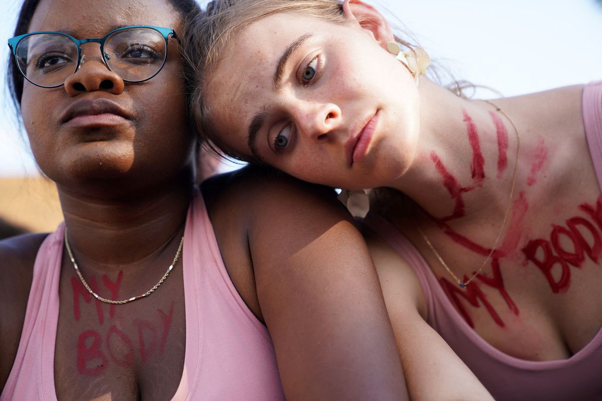 A Black woman and a white woman sit together on the steps of the Supreme Court, the white woman’s head resting on the Black woman’s shoulder. Both have the words “my body” painted on the skin of their chest.