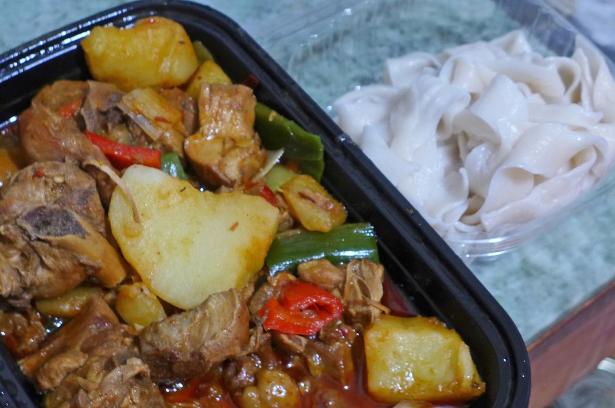 A plastic container of chicken and potatoes flecked with red bell peppers, with white broad noodles on the side.