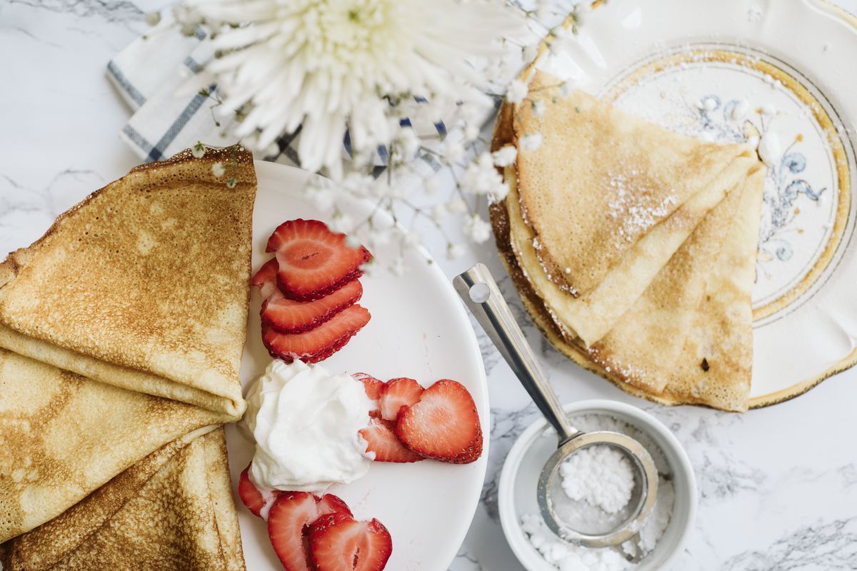 Two white plates of crepes, one with strawberries and whipped cream, on a marble countertop with a flower and a bowl of dusting sugar.