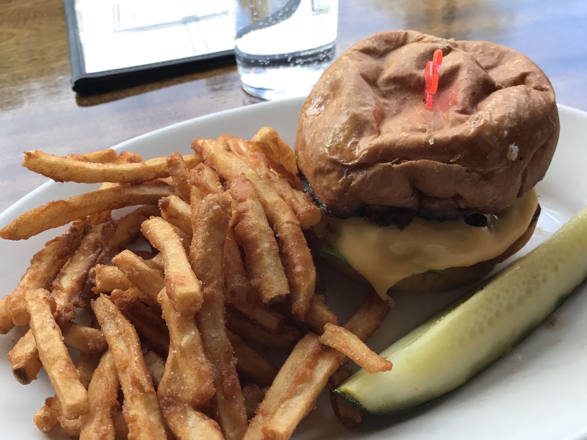 A cheeseburger, pickle wedge, and side of french fries are arranged on a plate. Behind them, a menu and a half-full glass of water sit on a countertop.