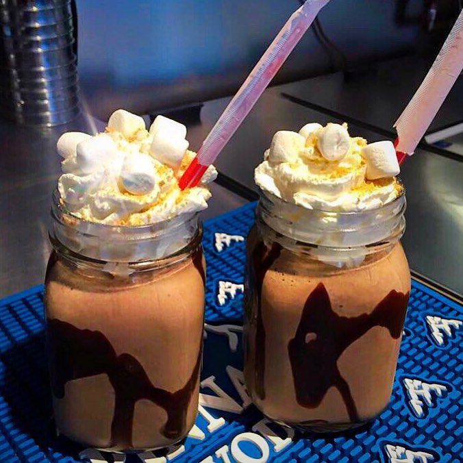 Two mason jars hold chocolate milkshakes with chocolate sauce, whipped cream, marshmallows, and big straws