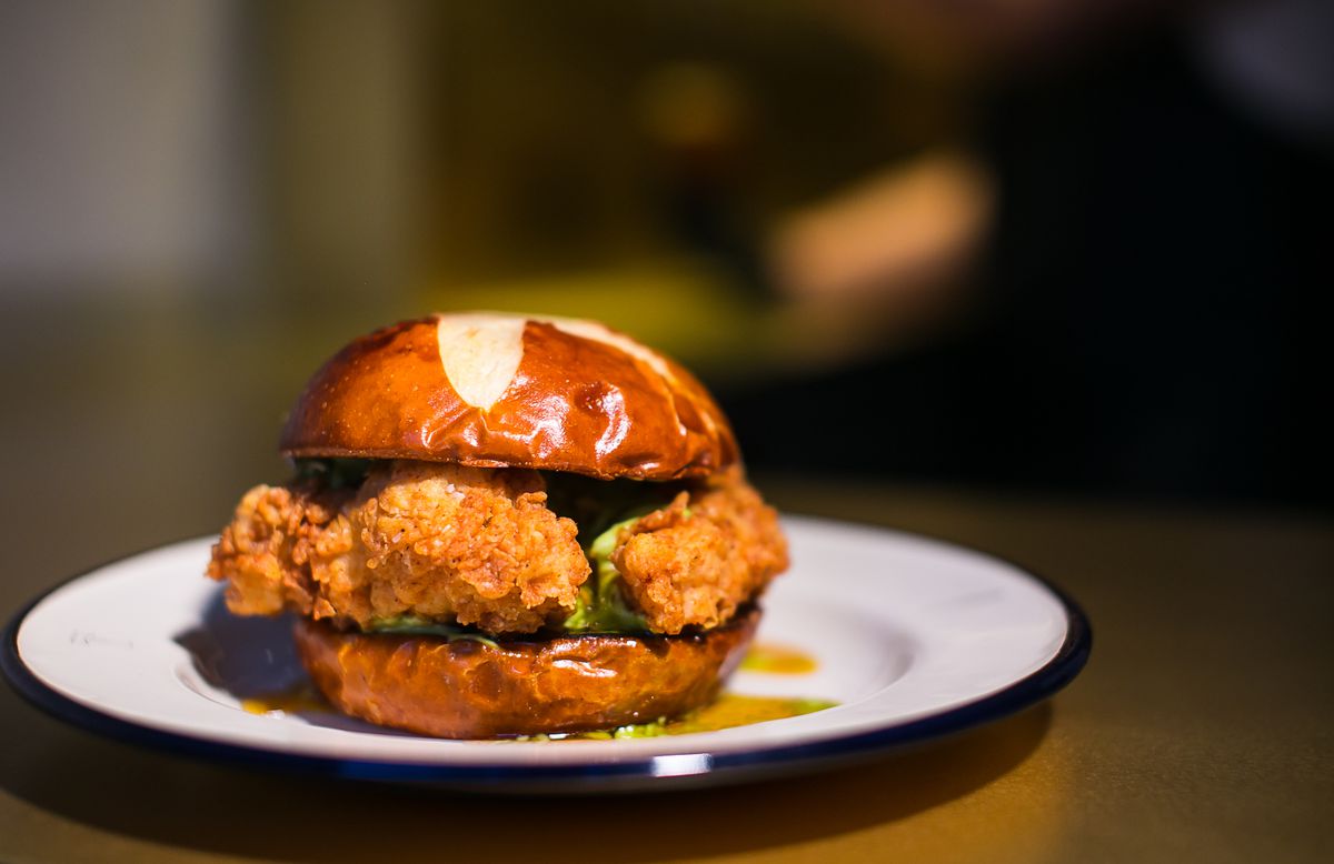 A crusty slab of fried chicken sits between two pretzel buns on a white porcelain plate