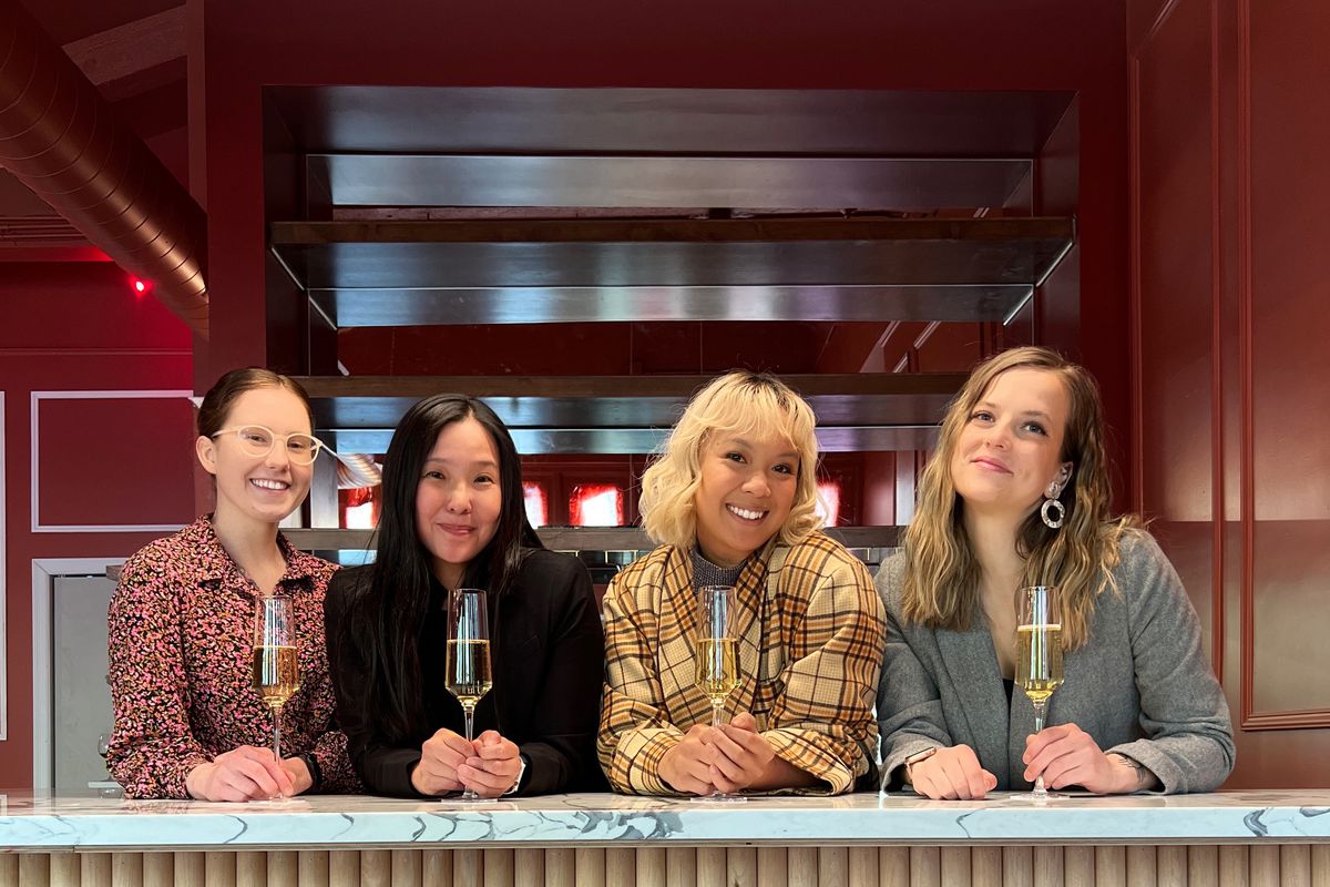 Four young women lean over a marble bar top, glasses of wine in hand.