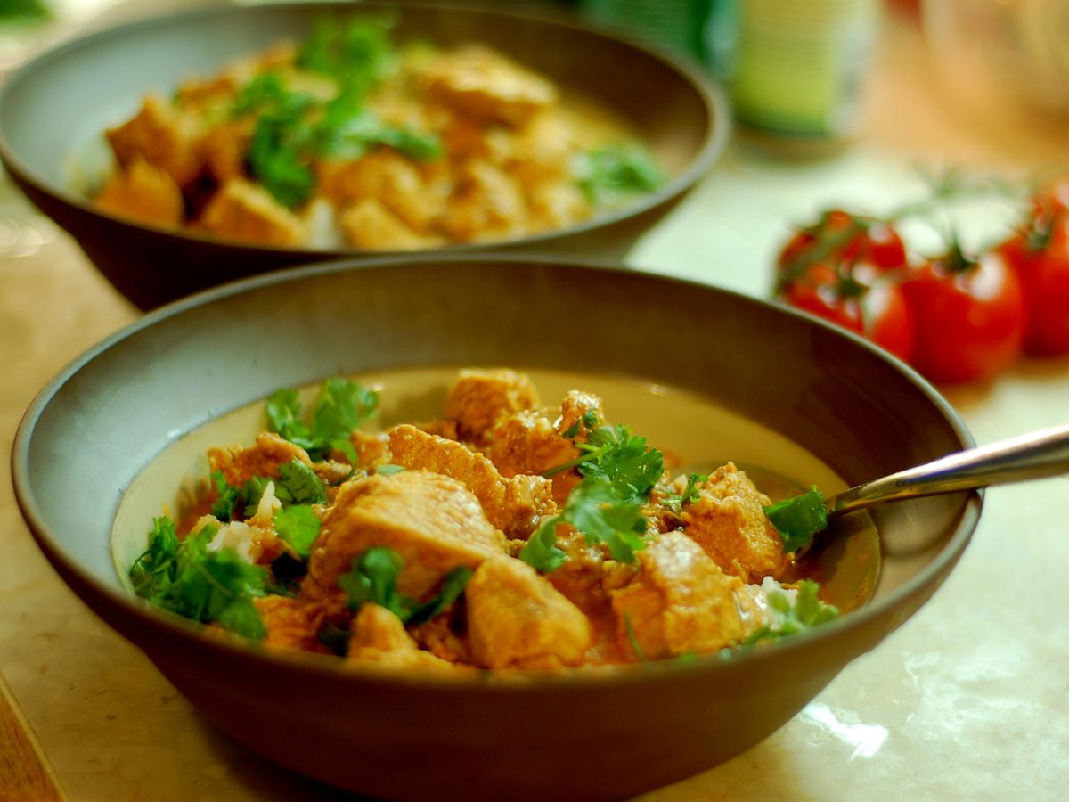 A bowl of tikka masala with chunks of chicken piled up in the sauce along with sprigs of cilantro, with another bowl and tomatoes on the vine in the background