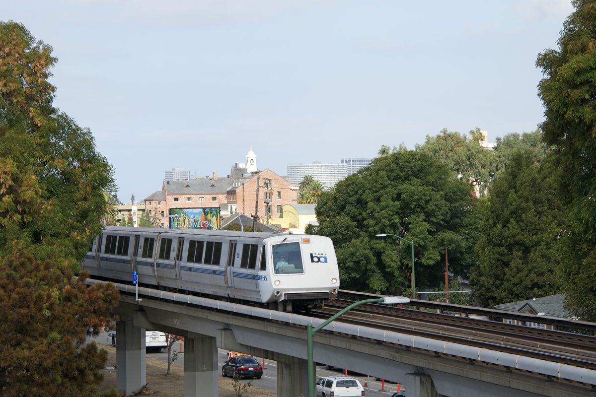 A white light-rail train on elevated tracks, with the tops of trees and buildings visible as it passes.
