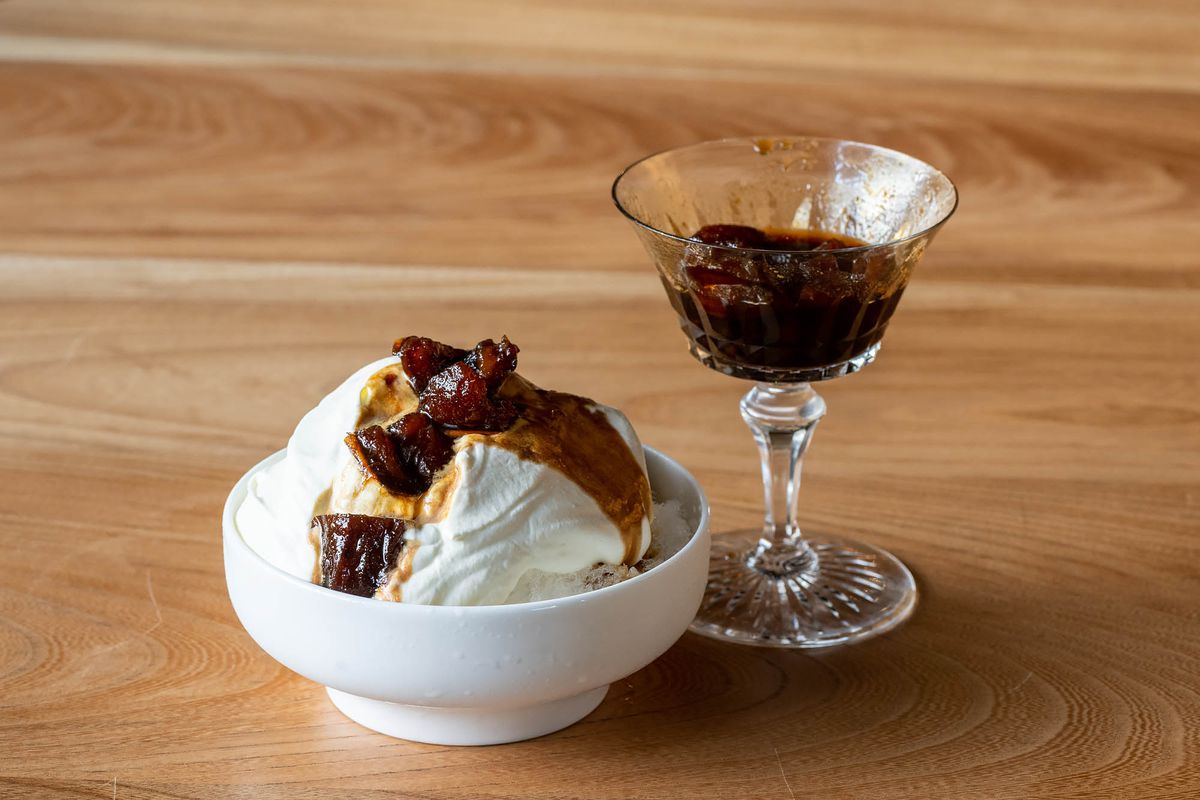 Dates and syrup pour down a white mound of ice cream in a bowl, with a holding glass beyond.