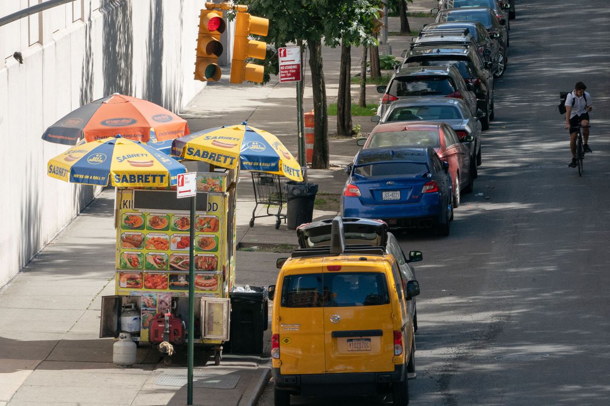 A brightly colored food cart with three umbrellas shading the cart from sun is parked on the sidewalk.