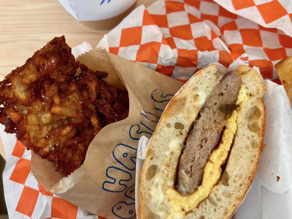 A sausage and egg breakfast sandwich next to a hash brown patty in a basket with orange and white checkered paper. 