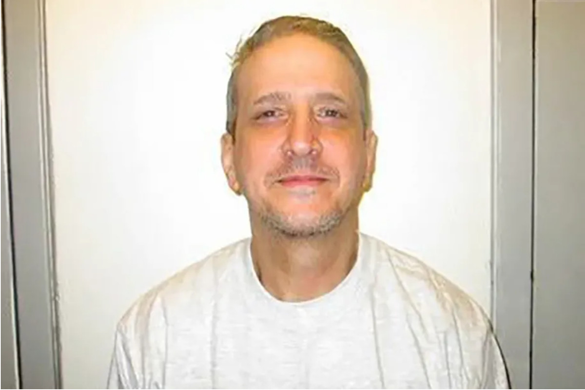 A white man with blond short hair in a white T-shirt smiles while looking at the camera, in a mug shot type photo.