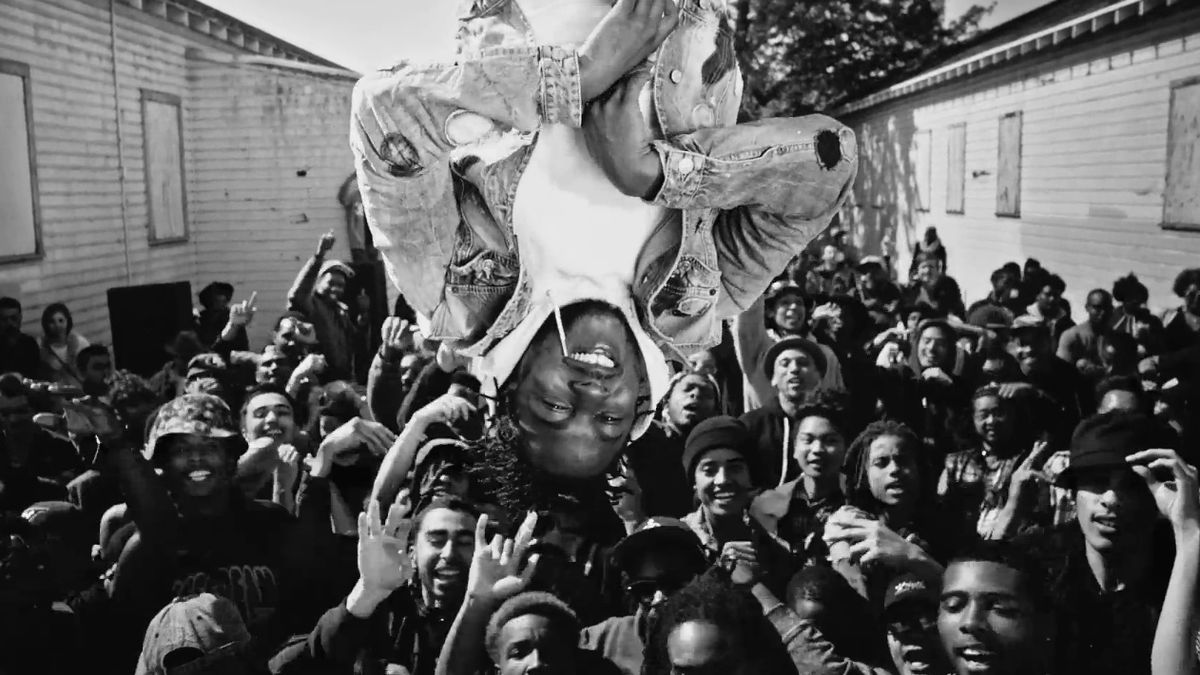 A man in a jean jacket (Kendrick Lamar) floats upside down over a crowd of people with their hands stretched upward, a visible smile on his face.