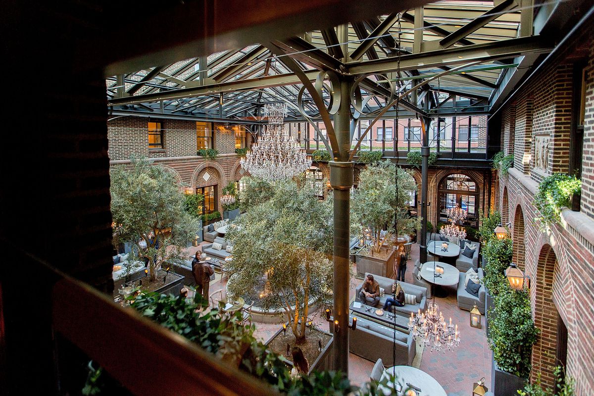 A gorgeous atrium, surrounded in brick walls and topped with a glass roof, filled with chandeliers, gray couches, and trees. 