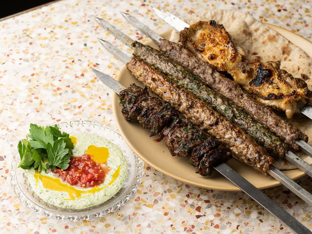 A wide shot of five metal skewers of grilled meat, plus bread, on a plate at a restaurant.