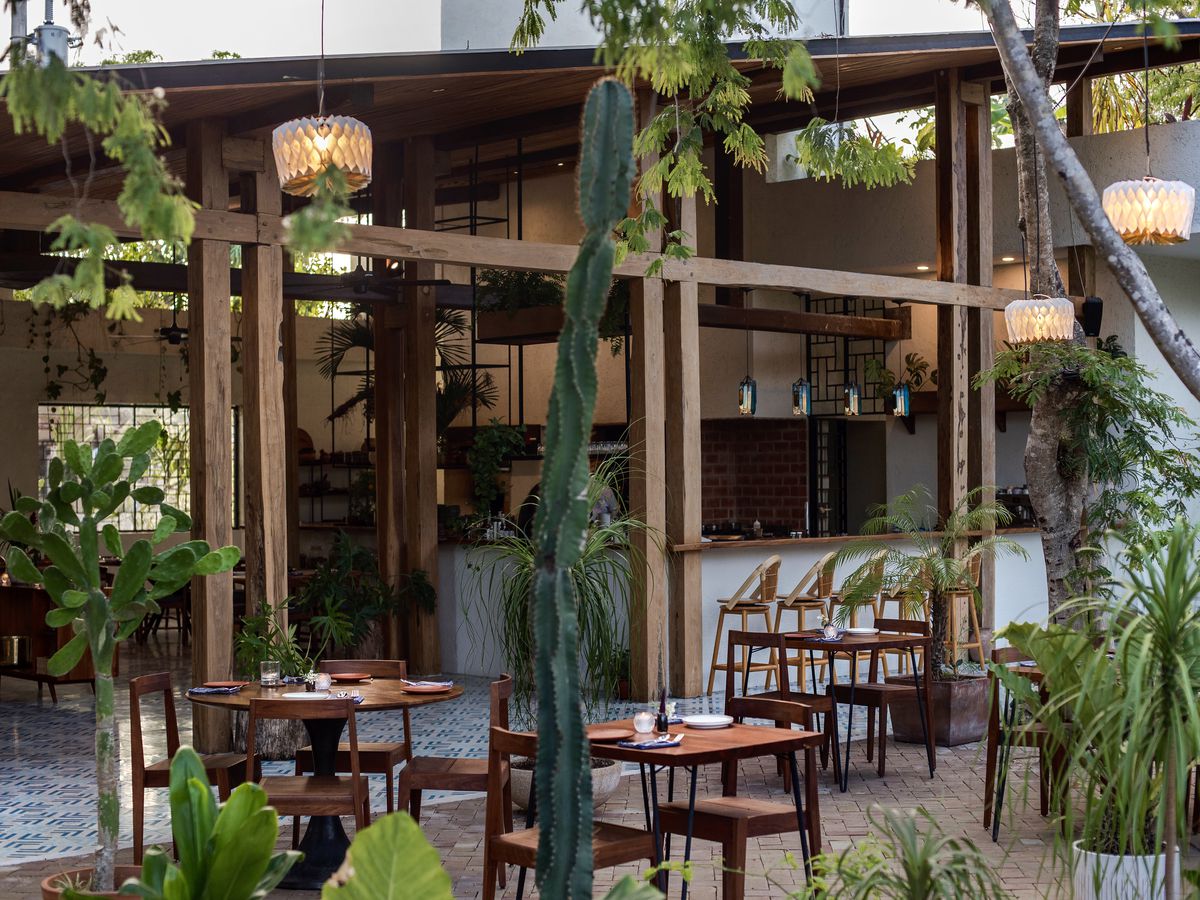 An open-air dining room blends seamlessly with an uncovered patio covered in potted plants and natural greenery. The space features long geometric wood beams, soft pendant lighting inside and out, tile flooring, and light wood chairs and bar stools.
