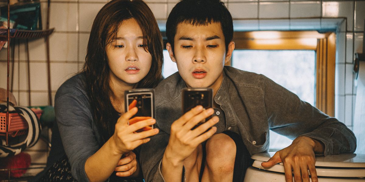 Park So-dam and Choi Woo-sik sit close to one another on the floor of a bathroom while each stares at their phone in the movie “Parasite.”