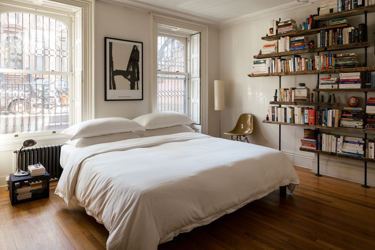 A bed is made with white linens. A shelf full of books is at right.