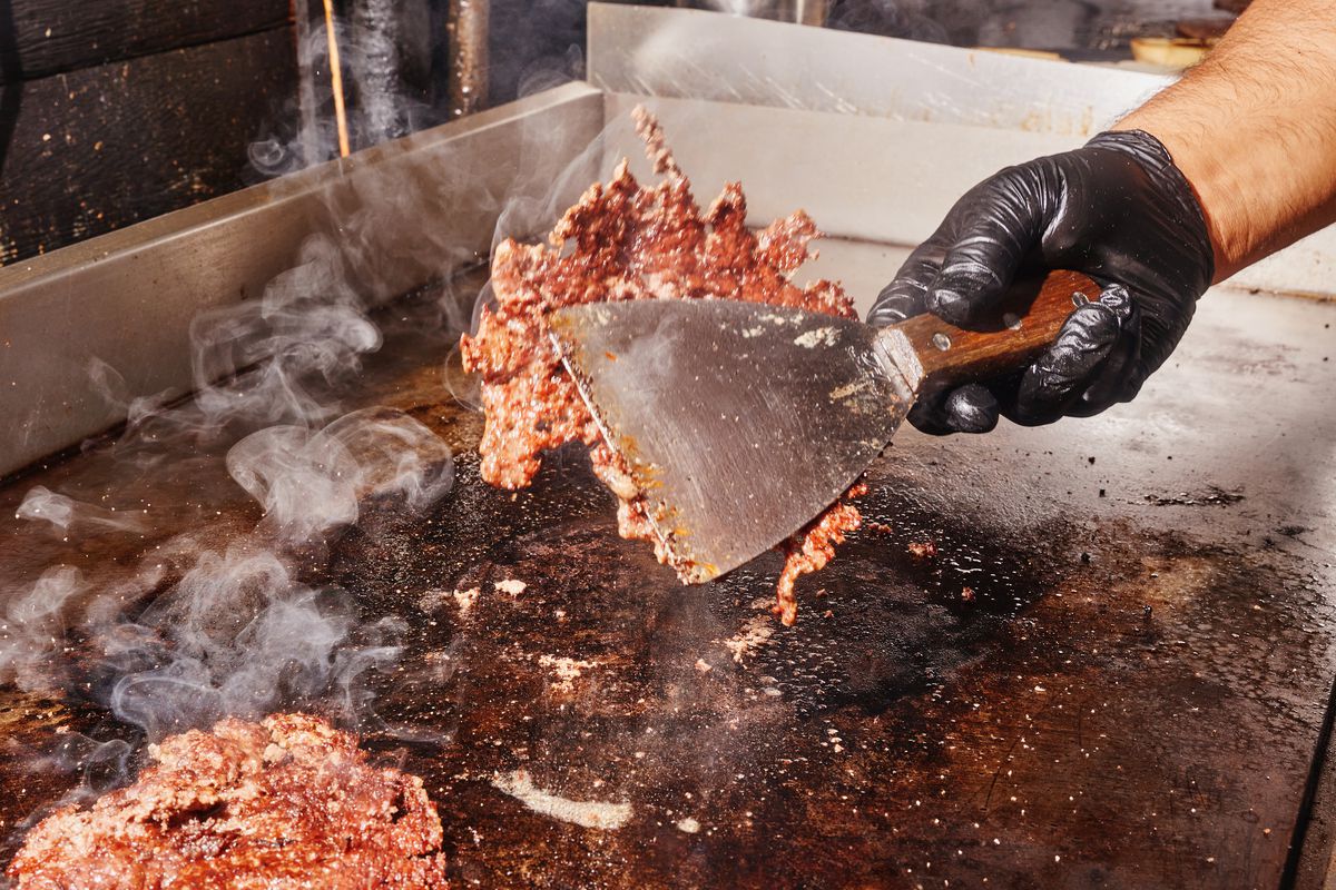 A hand wearing a black glove flips a lacey, paper-thin smash burger onto a griddle.