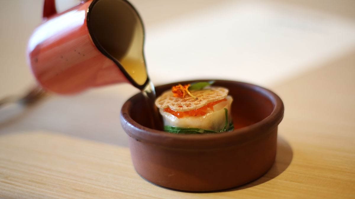 A small clay dish with a round morsel of walleye wrapped in sheeted sweet potato, topped with orange troute roe and a piece of wild rice tuile on a light wooden table. In the top left, a hand is pour a small pitcher of clear liquid into the dish.