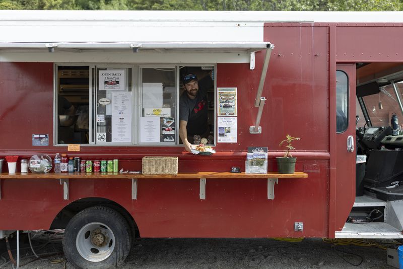 A red food truck with a man leaning out of the window wearing hat, presenting food. Canned and bottled drinks are lined up on a small wood counter in front of the truck.