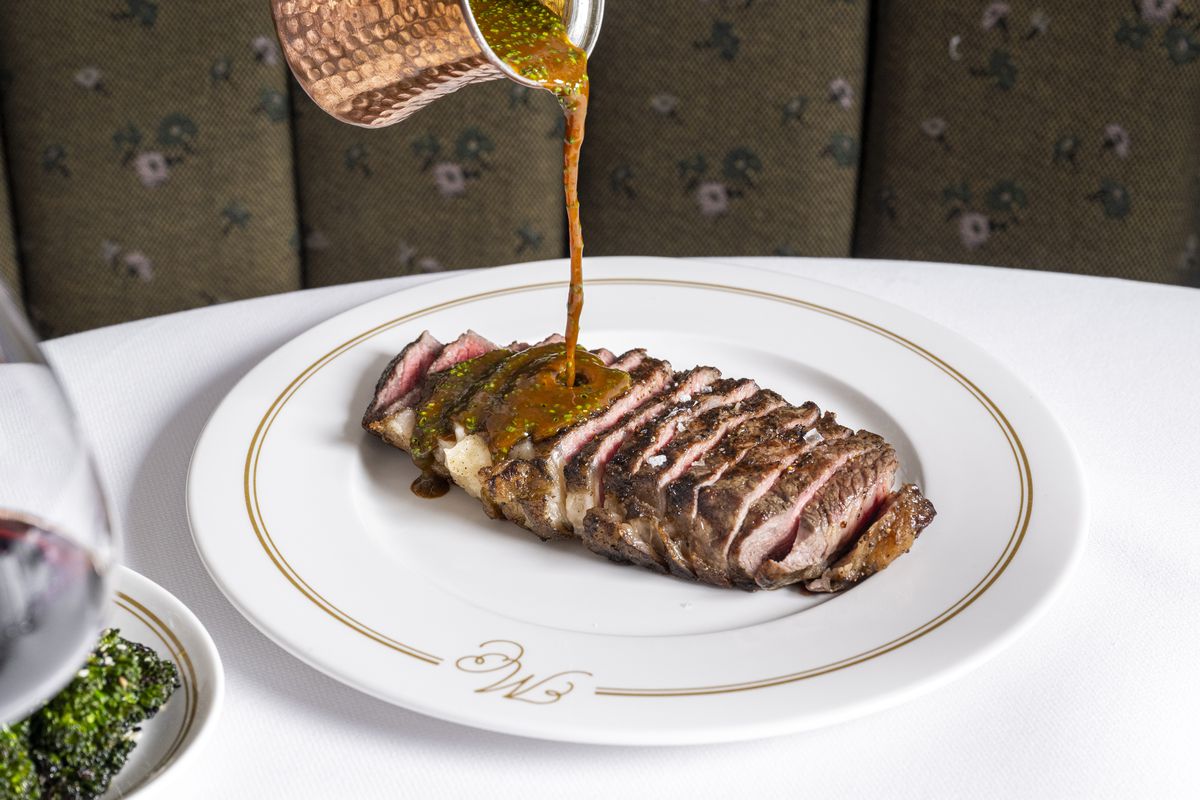 A sliced steak is served on a white platter, as a hand holds a copper container of au poivre sauce and pours it onto the steak.