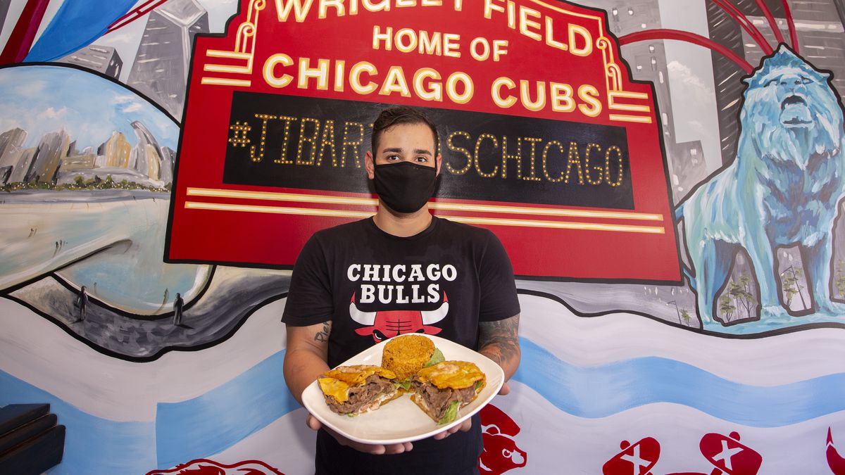 A man in a black mask and black Chicago Bulls t-shirt stands in front of a wall painted with a mural of the big red Wrigley Field sign holding a plate of jibaritos.