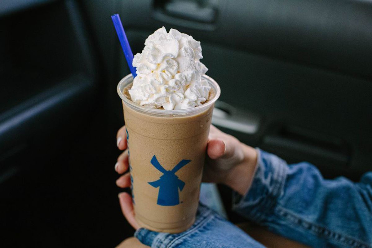 A white hand holds a plastic cup of a blended brown coffee drink, topped with a lot of whipped cream. A blue straw sticks from one side. The man is sitting in a car.