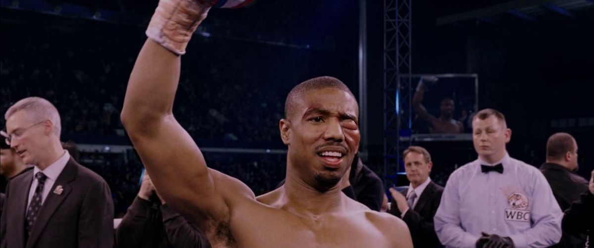 Michael B. Jordan as Adonis Creed lifting their fist up and smiling with a swollen left eye in Creed.