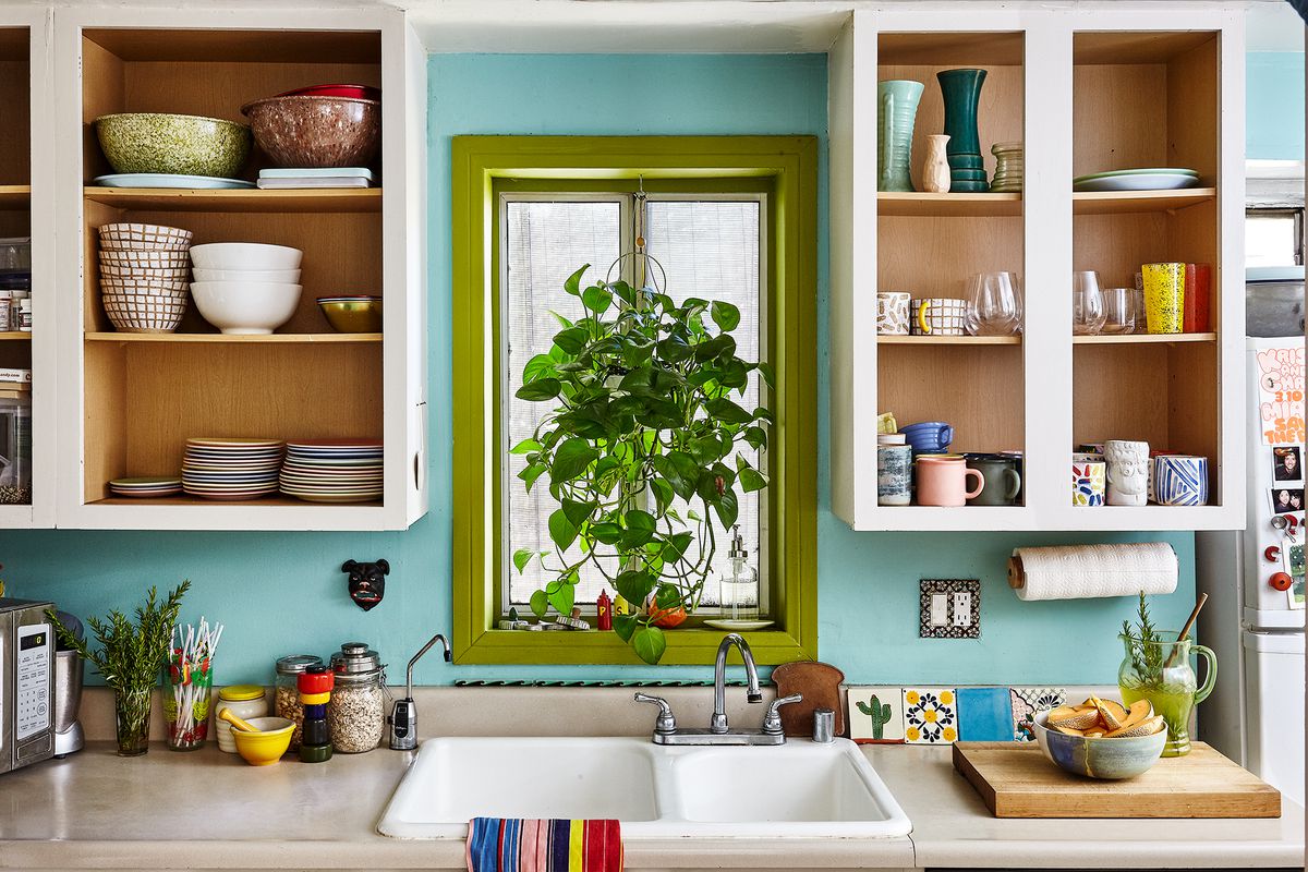 A kitchen with light blue wall and green window trimming. The window sits above the sink and between two white shelves filled with cups, bowls, and dishes.