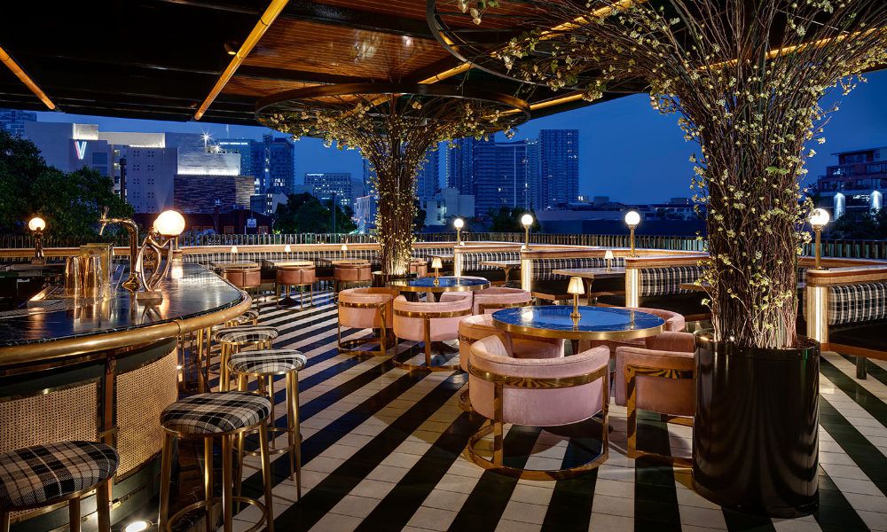 The roooftop patio at Born &amp; Raised is shown at nigh with columns made to look like trees with twinkling lights and a black and white striped floor. Chairs around circular tables have pink upholstery with brass legs. 