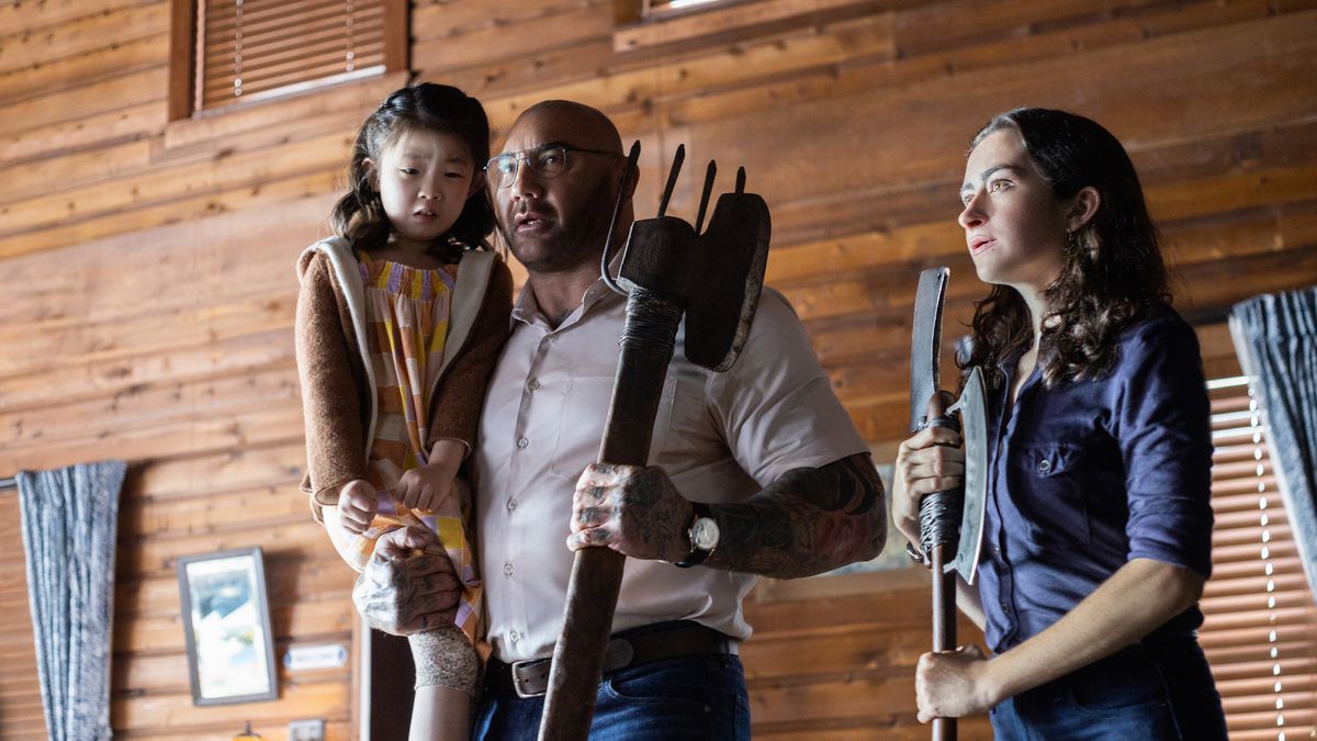 Home invader Leonard (Dave Bautista) holds weeping child Wen (Kristen Cui) in one hand and a pitchfork-like weapon in the other as his companion Adriane (Abby Quinn) clutches a butcher knife in M. Night Shyamalan’s Knock at the Cabin