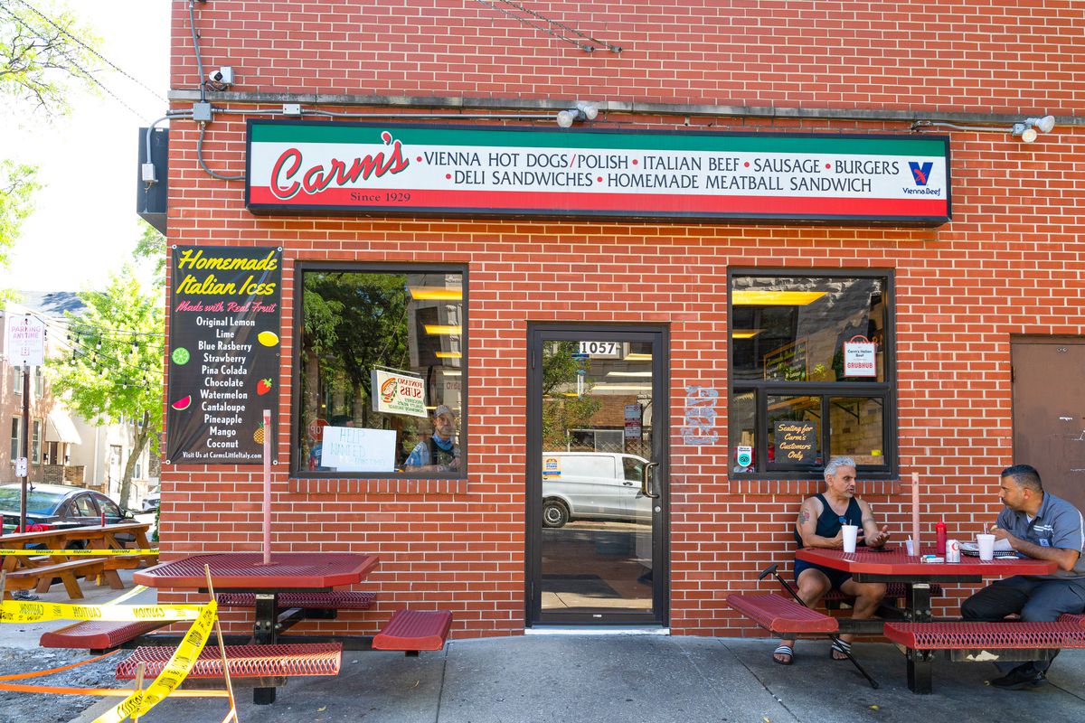 Two men sit at a red picnic table outside a red brick building with a sign that reads “Carm’s.”