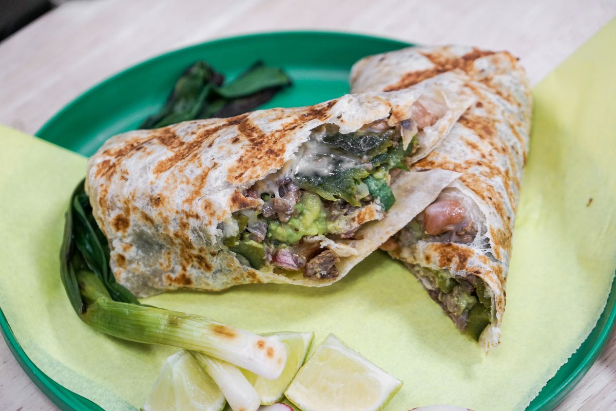 Grilled, split carne asada burrito with green onions on a parchment and green dish.