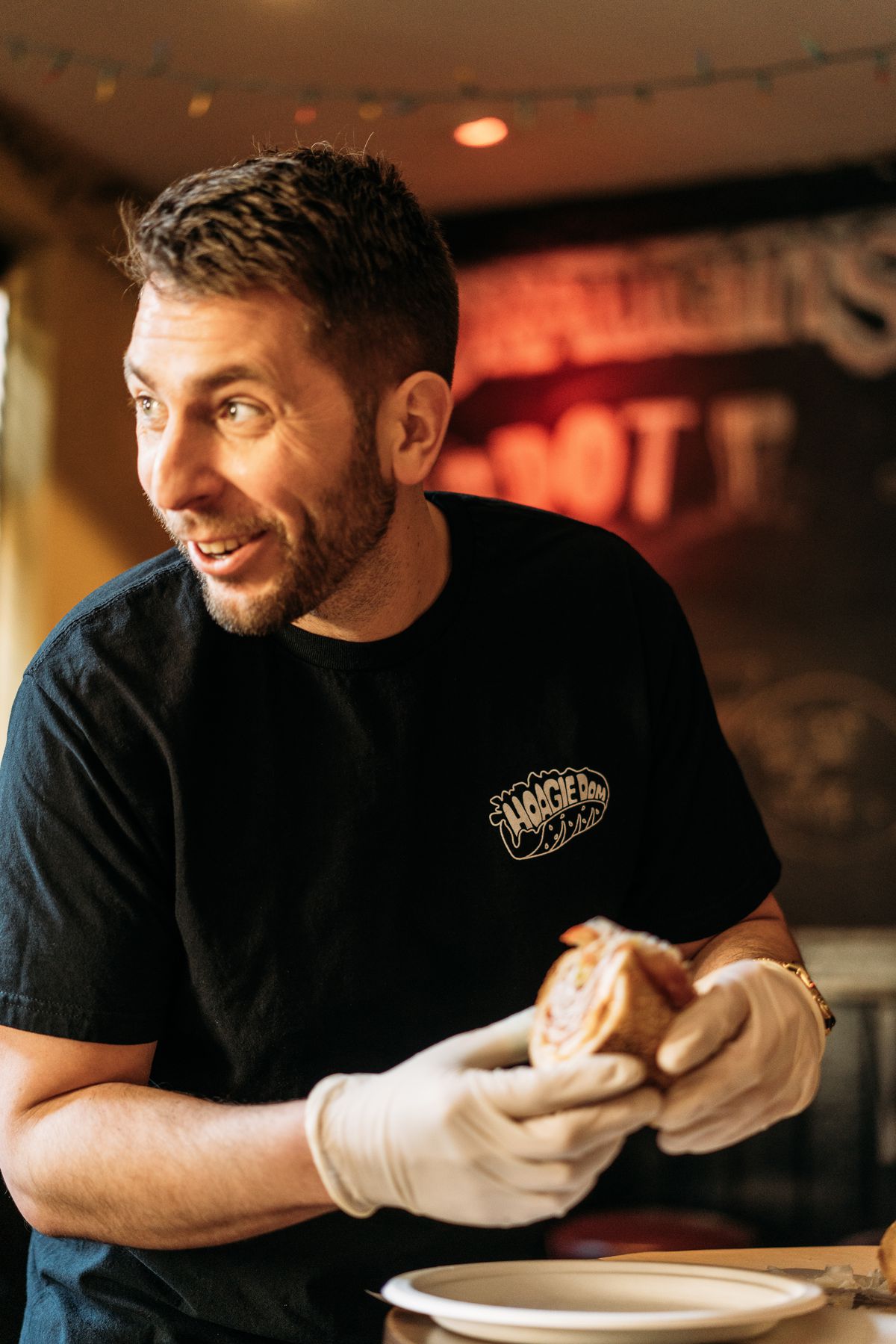 A man in a black T-shirt with a beard holds half of a hoagie in his hands while smiling to the left.