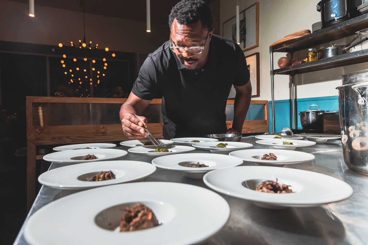 A Black man wearing a black shirt and clear glasses uses tweezers to place a piece of food onto one of several plates lined up on a table.