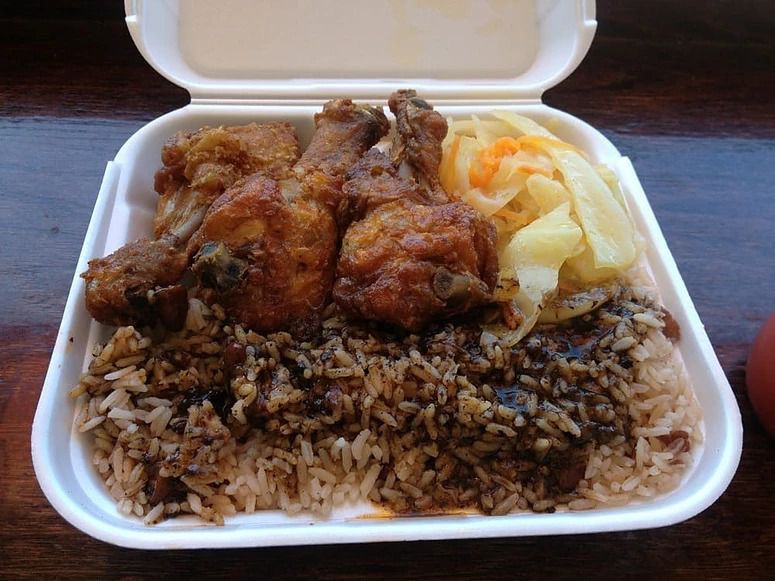 A white styrofoam takeout container is open to reveal Jamaican food, including rice and chicken drumsticks