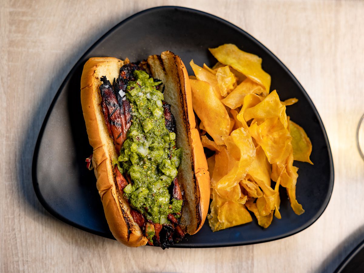 From above, an oblong plate holding a sandwich in a hotdog bun with a pile of potato chips. The sandwich consists of grilled strips of mushroom topped with chunky green sauce