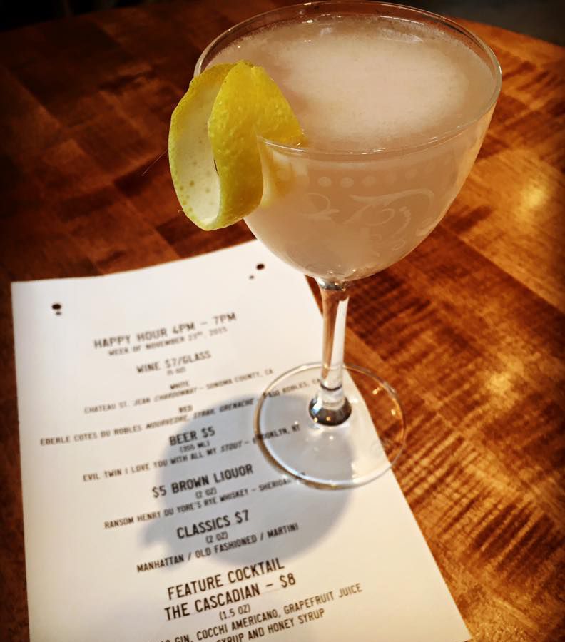 A frothy cocktail in a nick and nora glass garnished with a lemon strip, sitting on a paper menu