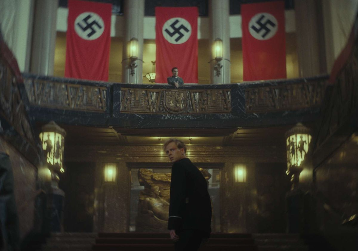 Wil, a young police officer with curly ginger hair, walks up stairs in a grand chamber decorated with Nazi flags. A German officer watches from a balcony