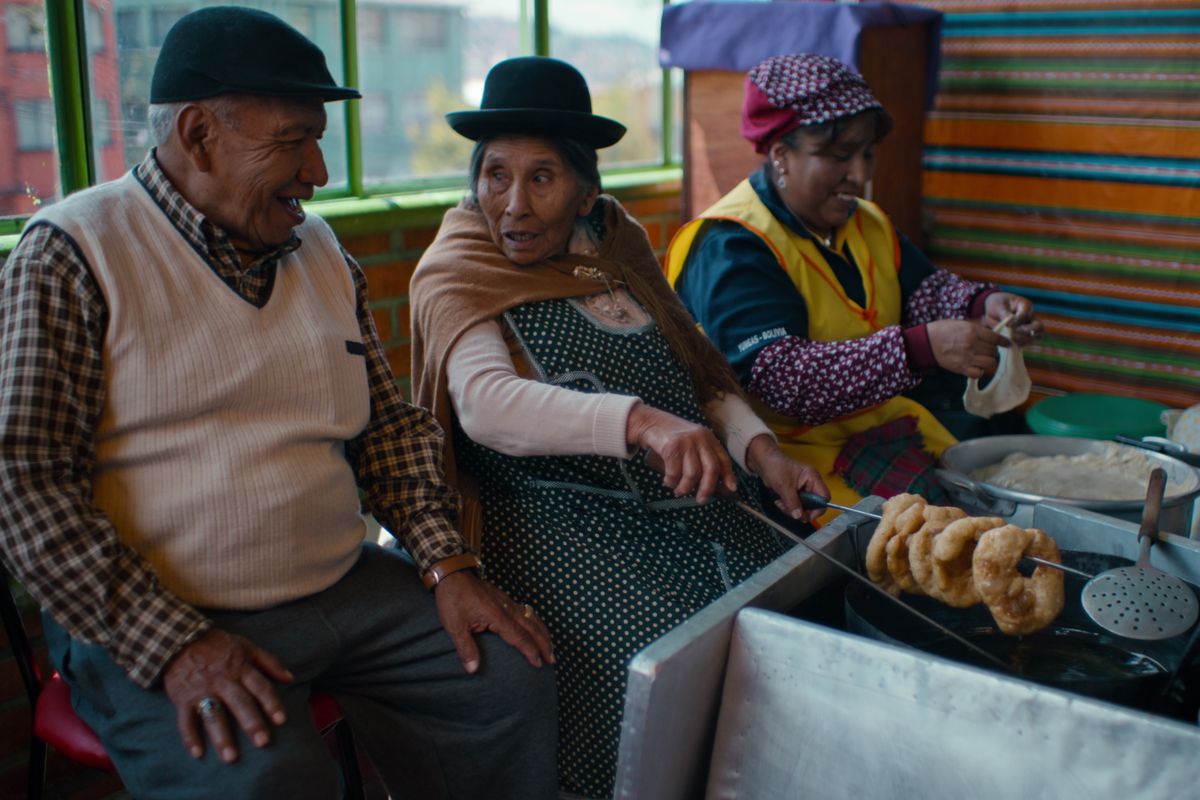 In Bolivia, an elderly man in a white sweater vest and black cap chats with two women in aprons as they prepare and fry dough in a room decorated in bright colored blankets.